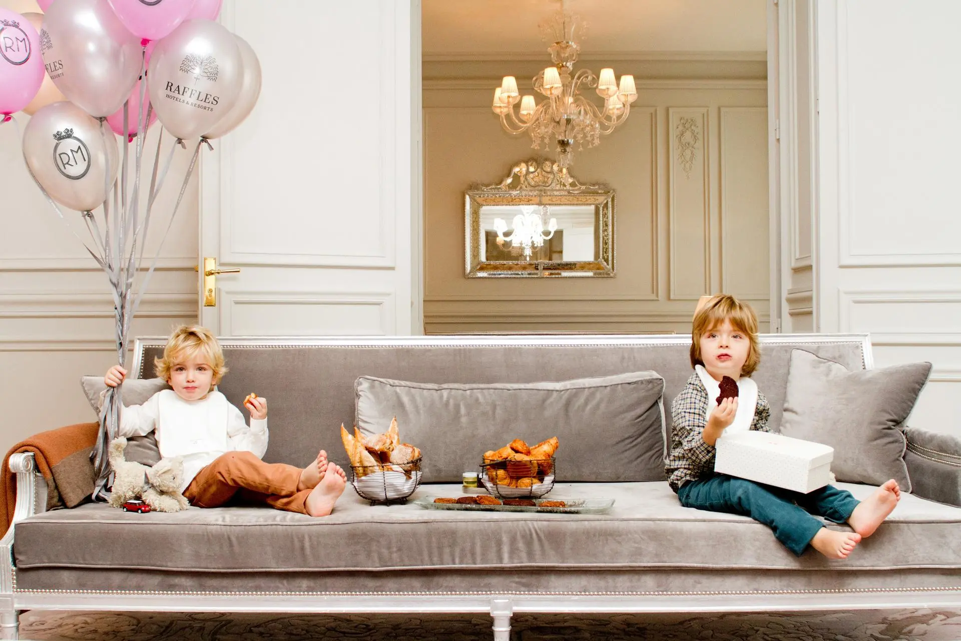 Two young children eating snacks on a luxurious grey velvet couch in a high-end hotel suite room, surrounded by branded Raffles balloons and pastries.