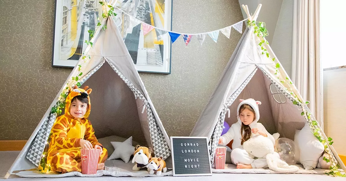 An ultra-luxury children's in-suite movie night teepee glamping setup at the Conrad London St. James hotel featuring two kids in animal onesies with popcorn.