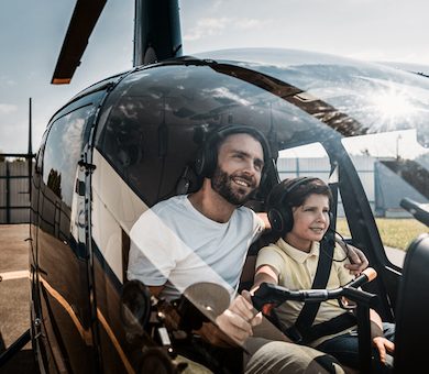 Portrait of happy unshaven father embracing glad boy while controlling helicopter with him. They sitting in cabin of it while talking. Optimistic parent and guy wearing earphones with microphones during sunny day