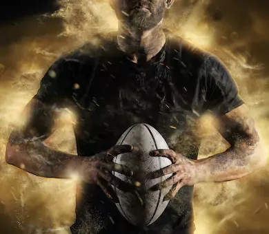 A muscular male athlete covered in dirt holding a rugby ball between both hands amid golden dust.