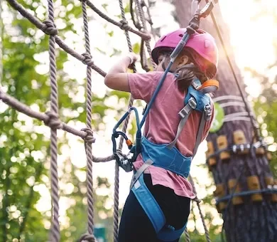 Young child wearing a safety harness and pink helmet navigating a high-altitude ropes course.