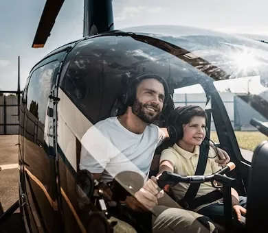A father and child wearing aviation headsets smiling inside the cockpit of a private helicopter over London.
