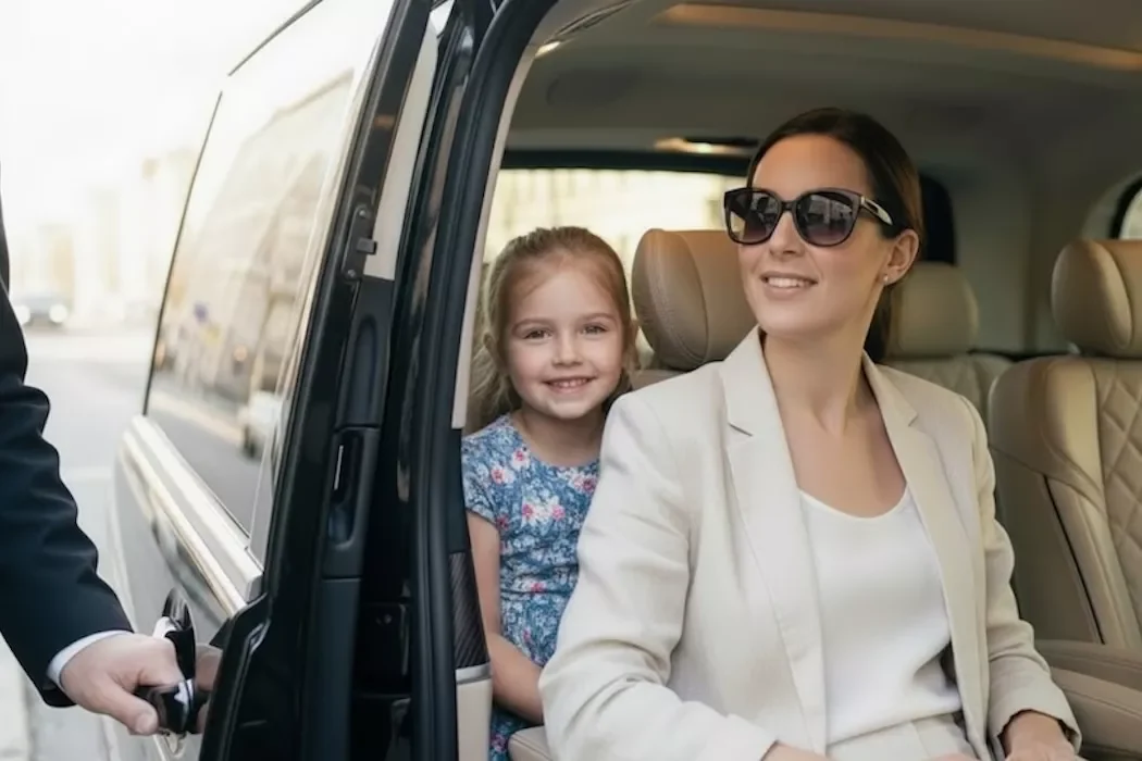 A smiling mother wearing sunglasses and her young daughter sitting in the backseat of a luxury chauffeur Mercedes V with the door open.