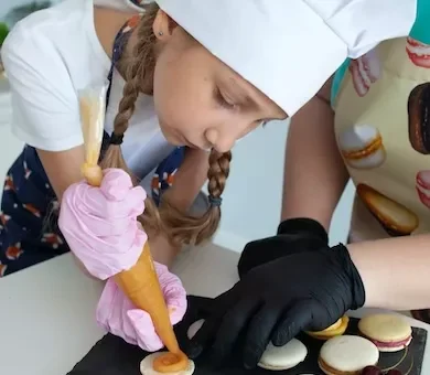 A young girl wearing a white chef hat piping colourful pink macaron shells during a private baking class in Paris.