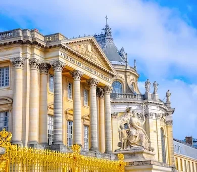 The magnificent golden gates and grand stone columns of the Palace of Versailles under a bright blue sky.
